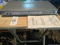 Silver Citizen DVD player front view with headphone jack, disc tray, and instruction manual placed beside it.