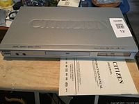 Front and top views of the silver Citizen DVD player model JDV-583-AA with visible front control buttons and headphone jacks, along with the instruction manual laid out in front.