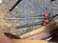 Photo showing all five long metal bar clamps placed on a gravel surface next to wooden decking and some black metal frames.
