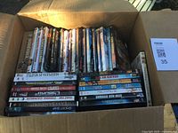 Box containing multiple DVD movies in various genres and titles showing spines and some front covers.