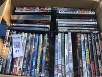 Photo showing a box of assorted DVDs stacked vertically and horizontally with visible titles in mixed condition.