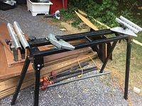 Metal saw stand shown outdoors on gravel surface, with adjustable roller supports visible on both ends for lumber support while cutting.