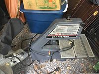 Side and front angle views of a gray Craftsman motorized band saw with 10-11 inch cutting capacity on a gravel floor, showing power cord and work table.
