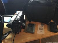 Sony Handycam camcorder powered on with battery pack, remote control, and carrying case visible on table.