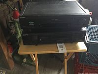 Image showing two black Sony electronic devices stacked on a small wooden table indoors. Top device is a VCR, bottom device is a tape player. Lighting is dim.