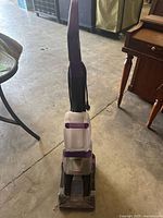 Front view of the upright Bissell carpet cleaner showing the handle and body with purple and gray coloring, standing on a concrete floor among furniture.