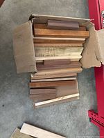 Open cardboard box filled with assorted wood blanks of various hardwood types including maple, walnut, cherry, and Red Oak, stacked neatly showing different colors and textures.