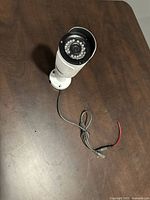 White wired bullet security camera with mounting bracket on a wooden table, cables visible without connectors attached.