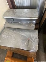 Three silver metal Enphase battery storage system enclosures stacked on a pallet, partially covered in plastic wrap.