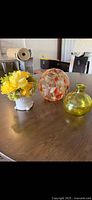 Three home decor tabletop accents on wooden table: yellow-green glass vase, clear decorative glass sphere with orange and white swirls, and artificial flower arrangement with yellow flowers in white ceramic pot.