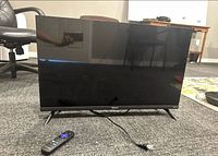 Front view of the Hisense 32-inch Roku smart TV sitting on floor with stand legs attached, power cable and remote placed in front, showing black screen and slim bezel.