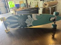Side view of the Kryptonics longboard skateboard with visible cartoon hand graphic on the deck and colored wheels.