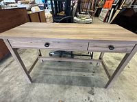 Full view of light brown wood grain desk on concrete floor. Shows two drawers and support stretcher below.