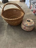 Two woven baskets side by side on a concrete floor: one natural tan handled basket and one smaller lidded basket with multicolor geometric design.