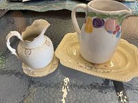 Two ceramic pitchers each on a matching dish. Small white and gold floral pitcher on scalloped dish; larger fruit motif pitcher on rectangular dish.