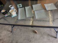 Seven assorted ceramic mugs in different designs and five textured clear glass plates arranged on a glass table.