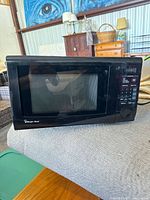 Black Magic Chef microwave oven placed on a cushioned surface showing full front and glass door.