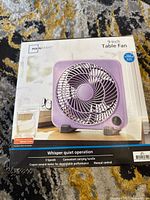 Photo of boxed Mainstays 9-inch violet table fan showing front of packaging with fan image and product features.