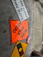 Collection of traffic and commercial signs including orange 'WORK AREA AHEAD', yellow traffic light sign, small Dairy Queen logo, and white 'ROAD CLOSED TO THRU TRAFFIC' sign on concrete floor.