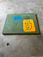 Front view of the abstract acrylic painting showing colorful abstract faces with botanical elements on green canvas.