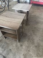 Two light brown wood grain composite end tables on a concrete floor. Rectangular shape with two shelves each.