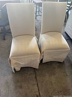 Pair of off-white upholstered Parsons chairs positioned side-by-side. Chairs have tall backs, cushioned seats, and skirted bases. Fabric shows moderate staining and wrinkling.