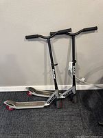 Two Razor kick scooters shown standing upright side by side against a wall on carpeted floor, with red wheels and black handles.