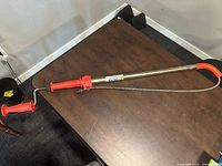 Full view of RIDGID handheld drain auger placed on a wooden table showing red ergonomic handles and metal shaft with coiled cable.
