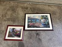 Two framed prints side by side, one larger landscape-oriented with a garden terrace scene, and one smaller cottage landscape scene with stone bridge, laid on concrete.