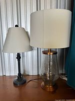 Two table lamps together, showing black metal base with cone white shade, and glass base with drum white shade