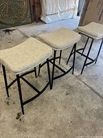 Three counter stools with black metal frame and light beige upholstered seats, side view showing shape and design.