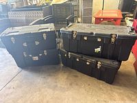 Four black plastic storage trunks stacked on the ground, showing exterior condition and metal latches.