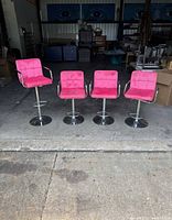 Four pink velvet bar stools with chrome metal legs and arms displayed in a row in a warehouse setting. One stool arm is broken.