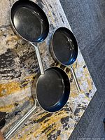 Three frying pans laid open showing black non-stick cooking surfaces and stainless steel handles on a patterned rug.