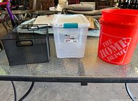 Set of three storage containers including black metal mesh bin, clear plastic bin with blue latch, and an orange Home Depot bucket placed on glass table.