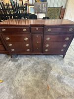 Front view of traditional dark brown wooden dresser with nine drawers, brass-tone oval pulls and tapered legs.