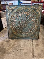 Full frontal photo of square turquoise metal decorative wall panel with raised floral circular pattern and distressed finish.