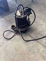 Side view of black submersible sump pump with attached electrical cord on concrete floor showing visible dirt and surface wear.