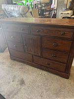View of wooden dresser showing 8 drawers and 1 cabinet door, medium brown finish, wooden and metal handles, with top surface wear visible.