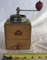 Front view of wooden coffee grinder showing label, hopper, crank, drawer and measuring tape for scale