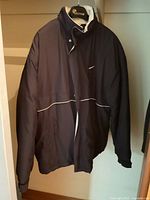 Front view of black Nike sport jacket on hanger showing swoosh logo, pockets, white piping