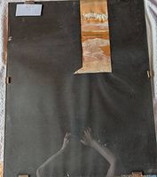 Front view of first framed artwork showing black mat board, central earth-tone collage, glass, metal clips