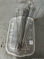 Set of assorted metal skewers placed inside a translucent plastic container on the ground, showing size and count.