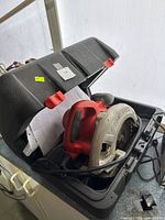 Circular saw with red and silver body inside a black plastic carrying case with red latches, visible saw blade and power cord.