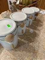 Four white ceramic cylindrical canisters with shiny white clamp lids and attached light wooden spoons arranged on a granite countertop with a slightly rustic background.