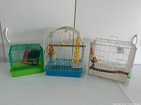 Full view of all three bird cages showing different sizes and colors: green/teal small cage, white dome cage with blue base, and white rectangular cage.