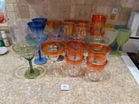 Overall collection of assorted colorful glassware showing various shapes like martini, margarita, tumbler, and goblet glasses in blue, orange, green, and clear with amber tones.