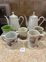 Front view of the two white ceramic teapots and five assorted mugs showing variety of designs including Steak & Ale mug and two-tone mug.