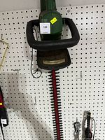 Photo showing full length of green electric hedge trimmer with dual black handles and red cutting blade teeth.