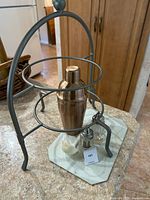 Front view of the metal stand holding the copper-tone cocktail shaker and other small bar tools on a countertop.